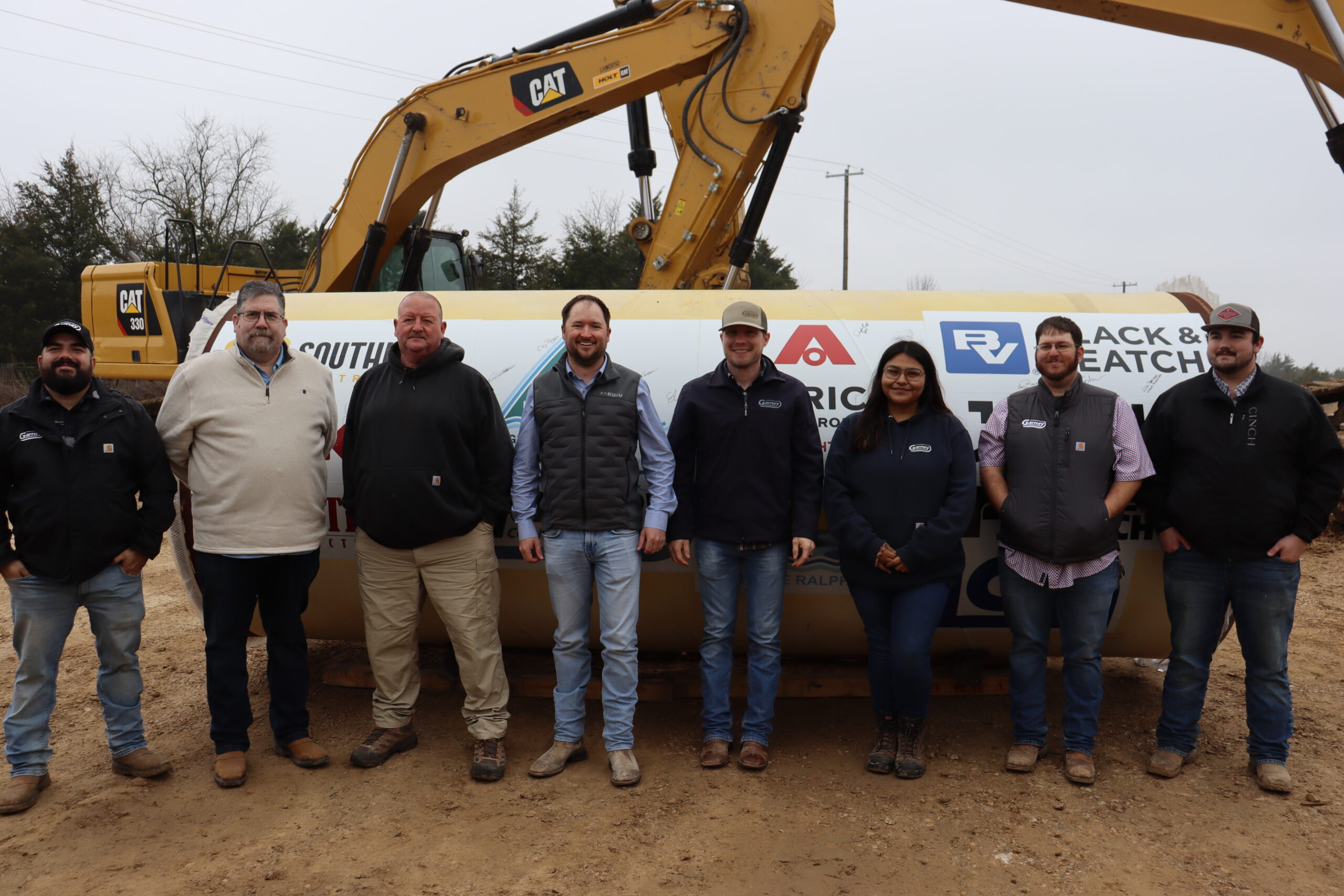 Upper Trinity team members, contractors and city/county officials pose in front of piece of pipe with logos of all project participants