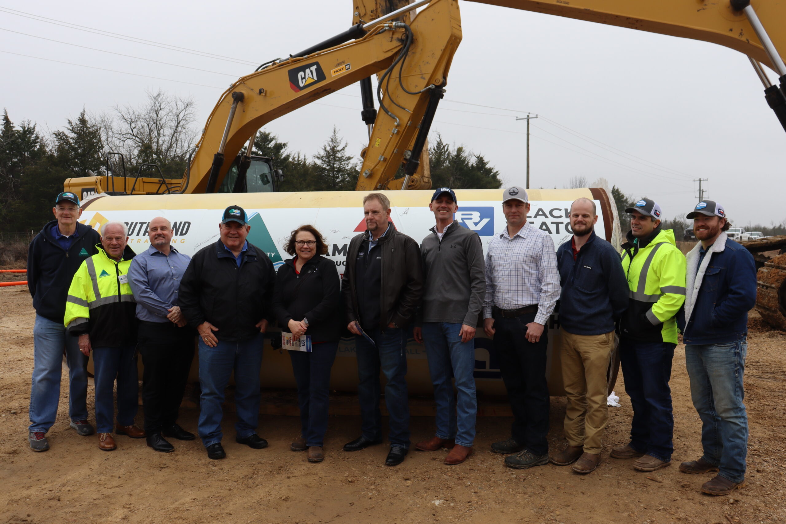 Upper Trinity team members, contractors and city/county officials pose in front of piece of pipe with logos of all project participants