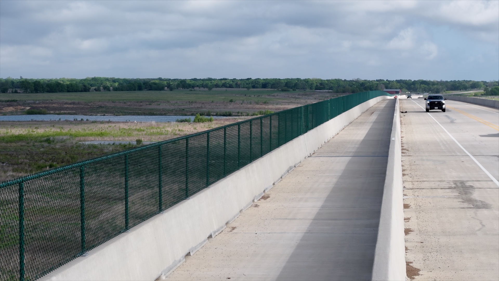Bridge roadway with concrete barriers and green metal fence, cars traveling on the right shoulder amid rural fields.