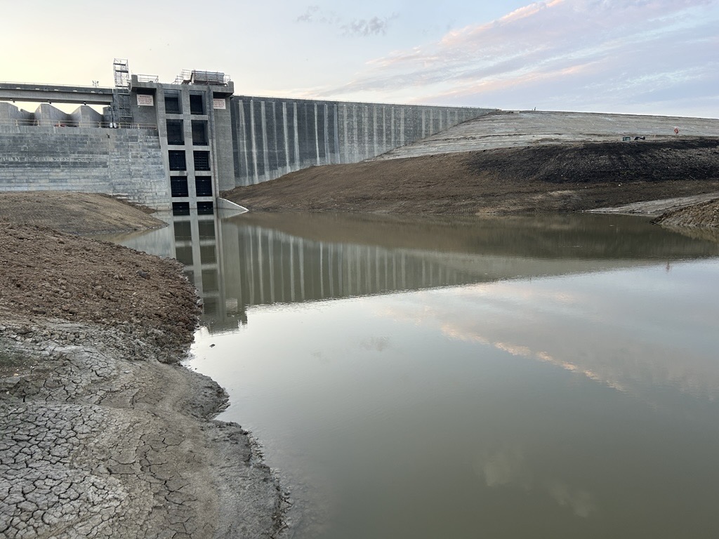 Large concrete dam with spillway gates, reflecting in calm water below.